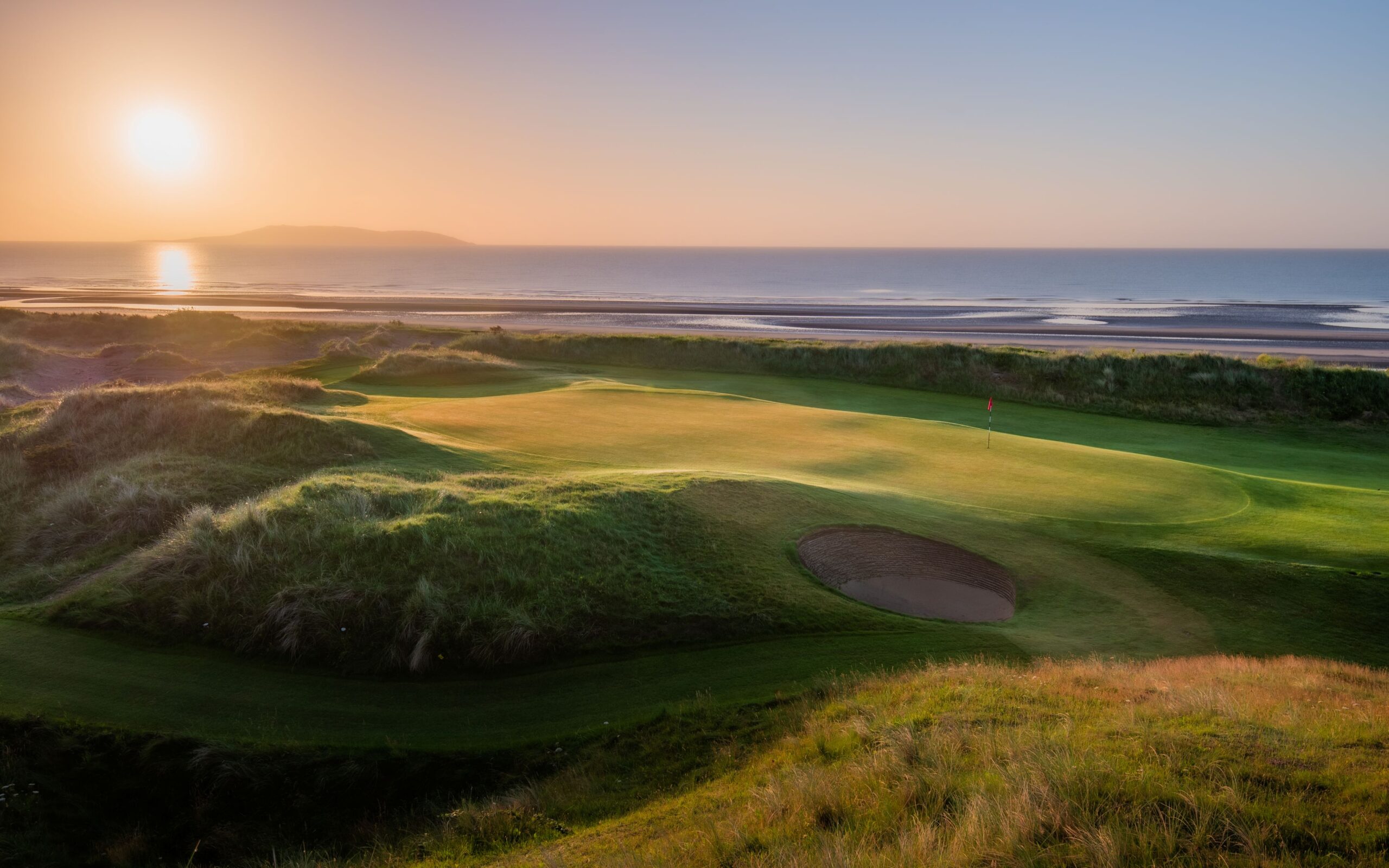 A stunning links golf course at sunrise, with a perfectly maintained green and bunker nestled among rolling sand dunes. The early morning sun reflects across the calm sea beyond the course, while golden light highlights the contours of the landscape, showcasing the dramatic coastal setting of The Island Golf Club.