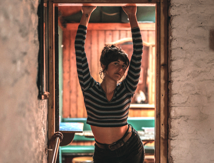 An individual with short dark hair leans forward in a doorway, bracing their hands against the frame. They wear a cropped, black-and-white striped long-sleeve shirt and dark trousers with a belt. Warm golden interior lighting contrasts with the cooler tones of the exterior textured wall.