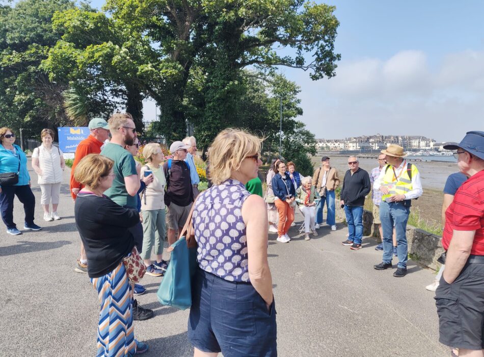 A sidewalk overlooking a coastal landscape with a tour group gathered along a stone wall.