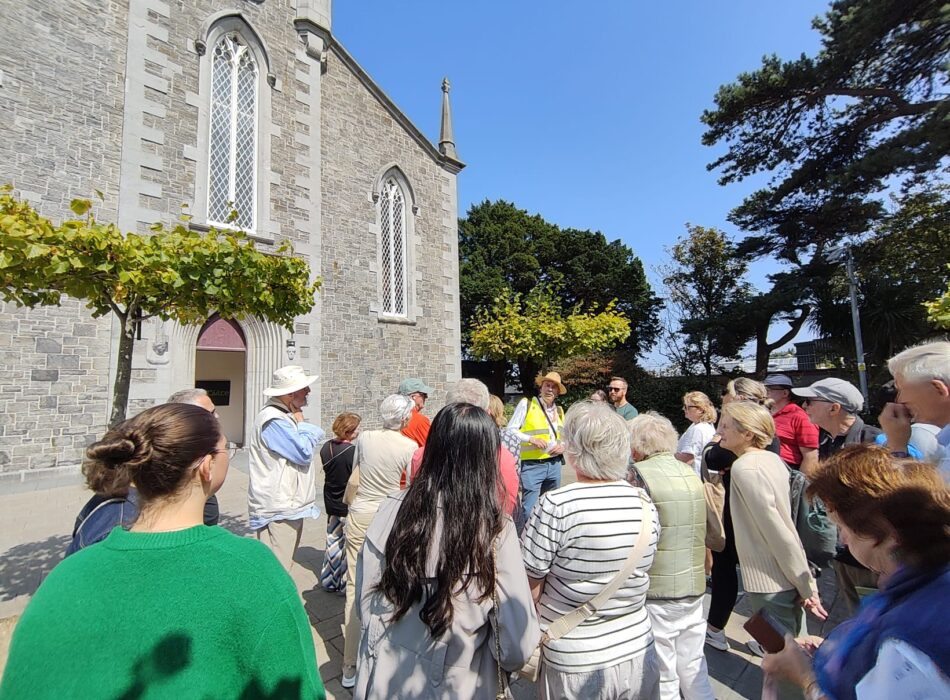 An outdoor photograph captures a tour guide speaking to a large group of tourists gathered outside a historic St Sylvester’s stone church under a bright blue sky.