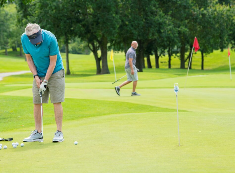 A daylight shot of a golfer wearing a turquoise polo, grey shorts, and a visor as he bends over his putter to practice on a smooth green. A cluster of white golf balls rests near his feet, and a small practice pin with a white marker stands a short distance away. In the soft-focused background, another golfer walks across the fairways among lush green trees, yellow pins, and red flags.