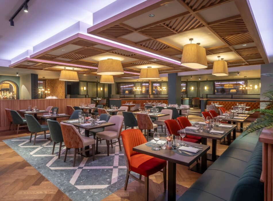 A large, open-concept dining room inside The Bracken Grill. The space is filled with dark wood dining tables meticulously set with wine glasses and cutlery. Seating includes a mix of plush armchairs in burnt orange, teal, and grey, alongside a dark teal tufted booth in the right foreground. The flooring features a striking combination of classic herringbone wood planks and a central section of teal and white geometric tiles. Overhead, a layered ceiling showcases decorative wooden slat panels, hanging drum lampshades, and soft purple cove lighting, while a bar area with arched windows is visible in the background.