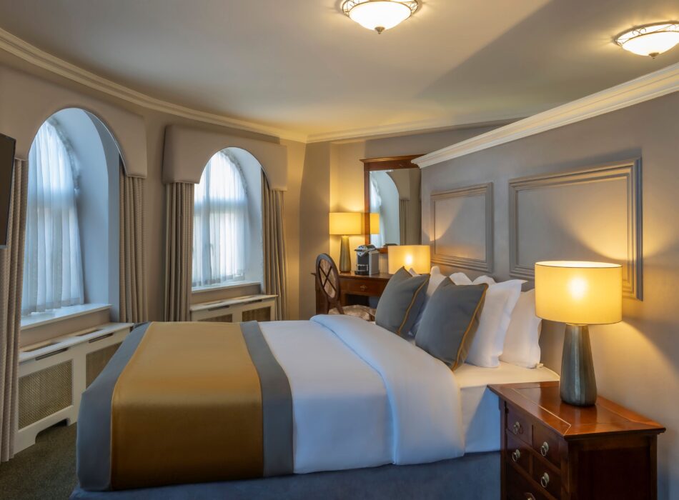 A beautifully decorated hotel room with a curved outer wall holding two tall arched windows dressed in beige valances and sheer curtains. The king-size bed is made with crisp white linens, grey accent pillows, and a satin gold and grey bed runner. The bed rests against a custom light grey partial partition wall designed with classic wood molding panels. A wooden nightstand with a glowing, warm lamp sits in the foreground, while a small vanity desk with a mirror and a coffee machine is tucked into the background corner.
