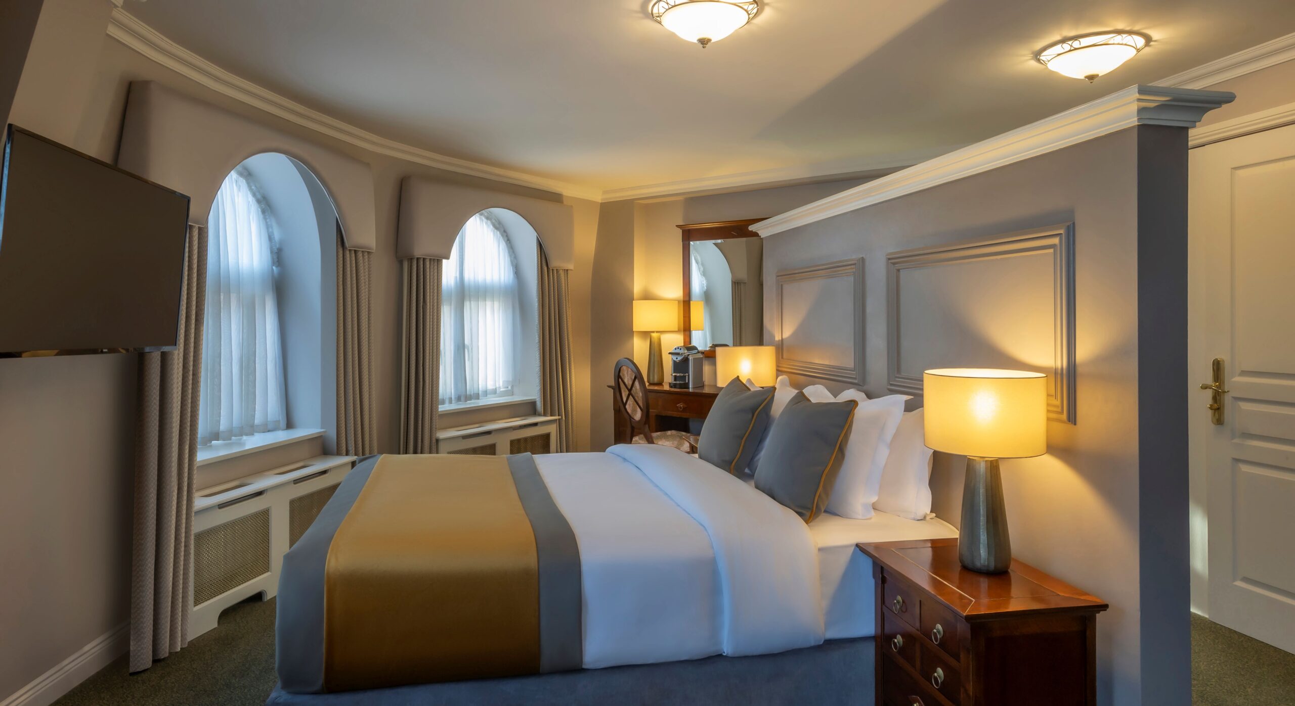 A beautifully decorated hotel room with a curved outer wall holding two tall arched windows dressed in beige valances and sheer curtains. The king-size bed is made with crisp white linens, grey accent pillows, and a satin gold and grey bed runner. The bed rests against a custom light grey partial partition wall designed with classic wood molding panels. A wooden nightstand with a glowing, warm lamp sits in the foreground, while a small vanity desk with a mirror and a coffee machine is tucked into the background corner.