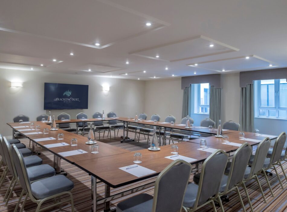 A spacious corporate meeting room configured with a large U-shaped wooden conference table and rows of grey upholstered chairs. Each seating station is neatly prepared with a white notepad, a pen, and a glass. Glass water bottles are spaced evenly across the table layout. The back wall features a large flat-screen television displaying the Bracken Court Hotel logo, flanked by two glowing wall sconces. Bright recessed spotlights illuminate the modern paneled ceiling, while two large windows with grey curtains line the right wall over a distinctive multi-colored striped carpet.