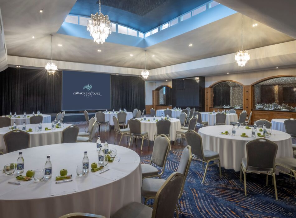 An expansive event ballroom configured with numerous round tables draped in crisp white tablecloths and paired with grey upholstered chairs. Each table is neatly set with writing pads, pens, bottled water, glasses, and fresh green apples. The room features a striking, multi-tiered ceiling with a large central skylight that reveals a deep blue upper section, illuminated by several glittering crystal chandeliers and recessed spotlights. A large projector screen at the far end displays the Bracken Court Hotel logo against a black curtain backdrop, and the floor is covered in a vibrant blue patterned carpet.