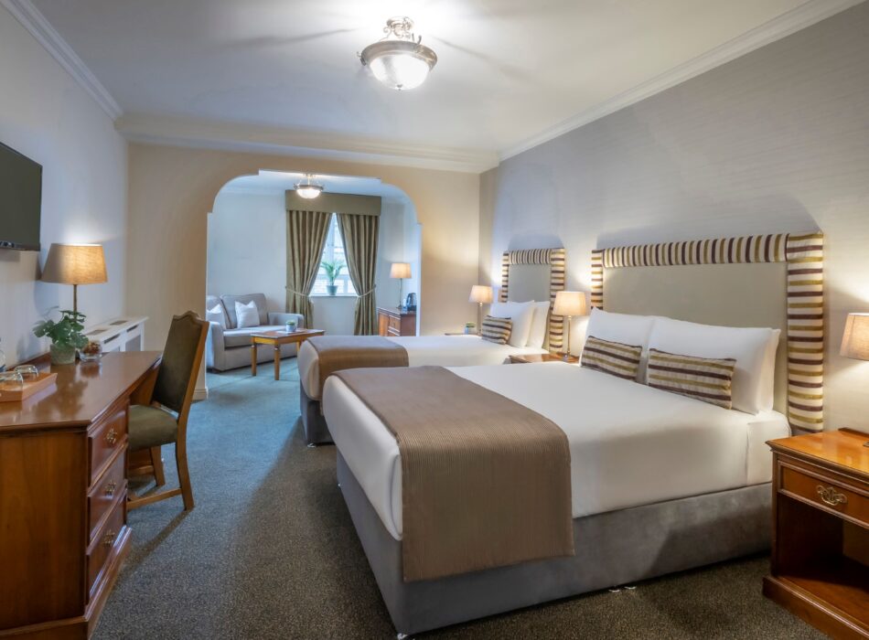 Spacious hotel family room with two large beds, warm neutral décor, bedside lighting, a desk and a separate seating area with sofa and coffee table.