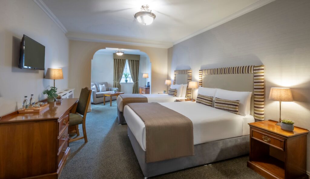 A well-lit, traditional hotel room showcasing two double beds dressed in crisp white linens, striped accent pillows, and bronze-beige bed runners. Elegant fabric headboards with matching striped borders frame each bed, flanked by dark wood bedside tables with classic lamps. On the left wall, a long wooden desk holds water bottles and a small plant beneath a wall-mounted TV. An archway in the background opens up into a separate sitting lounge equipped with a grey sofa, a coffee table, and curtains framing a window.