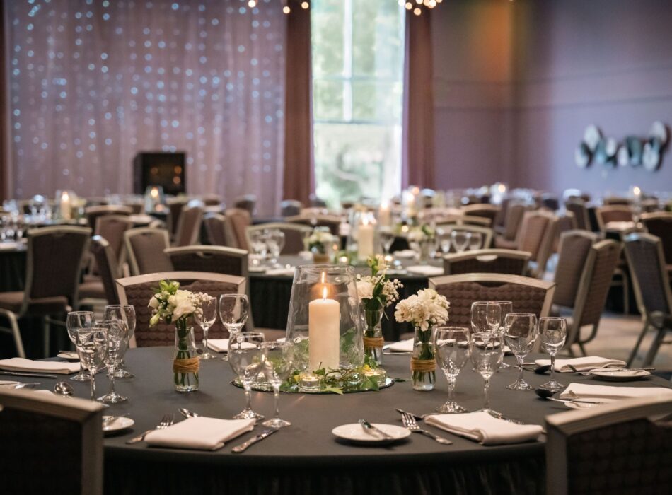 A large ballroom or event space prepared for a formal reception. The foreground features a round table draped in a dark tablecloth, meticulously set with silver cutlery, white folded napkins, and multiple wine glasses. The centerpiece consists of a large, lit white pillar candle inside a glass vase, surrounded by small glass jars holding delicate white flowers. In the softly blurred background, numerous other banquet tables and fabric-backed chairs fill the room. A sheer curtain illuminated by fairy lights serves as a backdrop on the left wall under warm, ambient lighting.