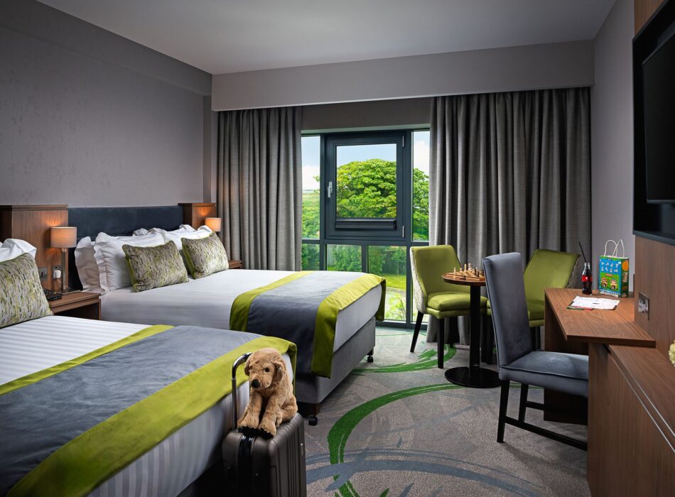 A modern twin hotel room featuring two beds with white linens, grey throws, and bright lime-green accents, a small plush dog toy sitting on a suitcase, a wooden desk with a grey chair, and a small seating area near a window showing lush green trees outside.