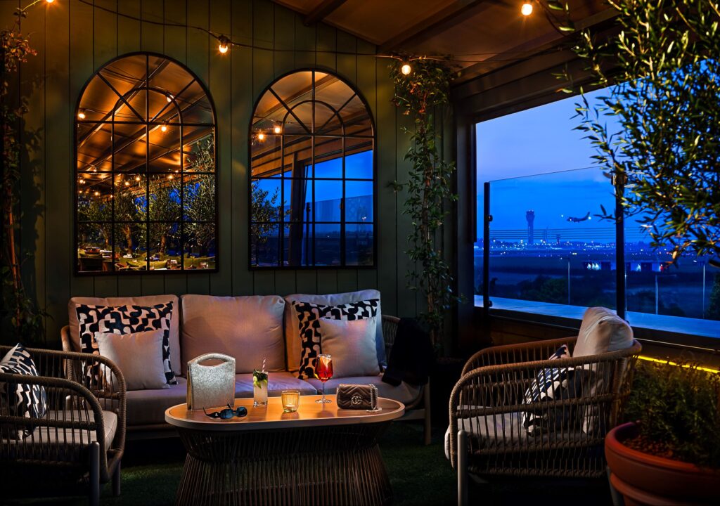 A modern outdoor terrace lounge at the Runway 28 Rooftop Bar & Restaurant. The space is furnished with a grey sofa and matching armchairs with patterned accent pillows around an oval coffee table holding cocktails and accessories. String globe lights illuminate a green panelled wall decorated with two large, arched multi-pane mirrors reflecting the interior. A large open window offers a clear view of an airport runway against a deep blue twilight sky, with an air plane in mid-flight and an air traffic control tower visible in the distance.