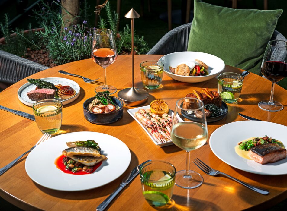 An overhead, sunlit view of a round wooden dining table beautifully set with a gourmet spread of various dishes, including seared fish, grilled prawns, steak, and a small dessert plate. The table is neatly arranged with silverware, multiple glasses of white, red, and rosé wine, ribbed green water glasses with lime slices, and a sleek, minimalist cordless table lamp in the center.
