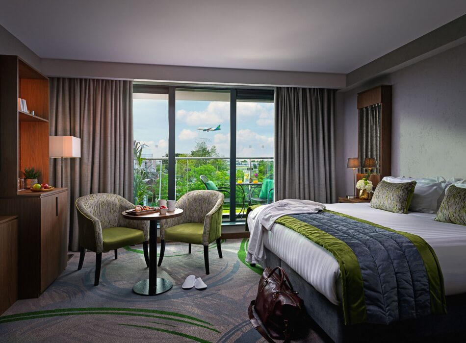 A contemporary hotel room featuring a king bed with white linens and a green-accented grey runner, a brown leather travel bag on the patterned carpet, a small round dining table between two green armchairs, and floor-to-ceiling windows leading to a balcony where an airplane is visible in flight through the glass.