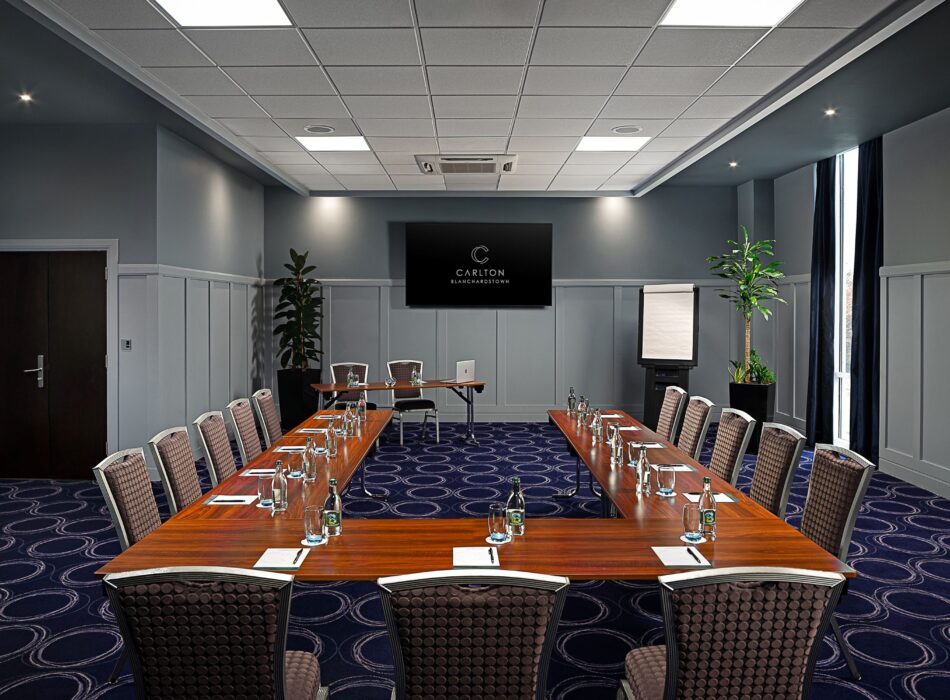 A modern hotel meeting room set up with a U-shaped conference table, chairs, and a presentation screen.