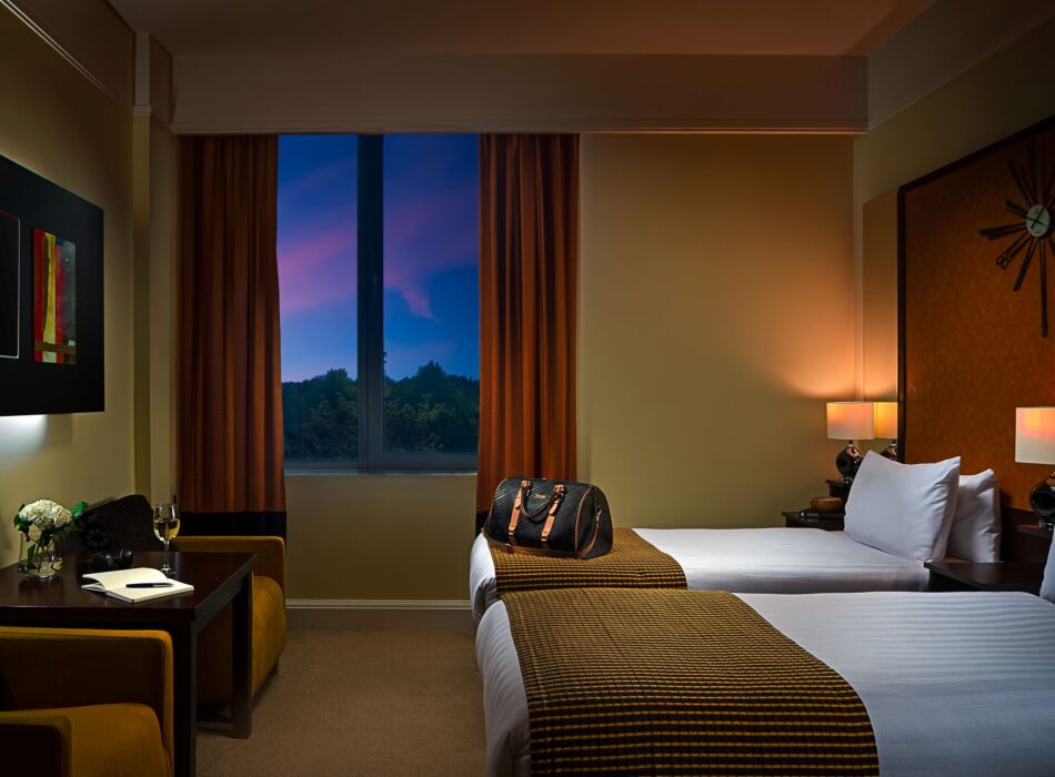 A dimly lit, warm-toned hotel room configuration with two twin beds made with white linens and textured bronze-brown bed runners. A black duffel bag rests on the far bed. Soft light radiates from lamps on a dark wood headboard wall decorated with a large mid-century starburst clock. To the left, a small wooden table sits between two armchairs, holding a wine glass and an open book. A large window in the center showcases a vibrant twilight sky with shades of pink, purple, and blue over a silhouette of distant trees.