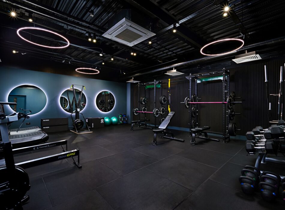 A modern, dimly lit indoor gym featuring a dark color scheme with industrial ceilings, illuminated by circular overhead ring lights. The facility is equipped with a Concept2 rowing machine, a treadmill, weight benches, two large power racks with barbells, a rack of dumbbells, kettlebells, and exercise balls arranged against a back wall with backlit circular mirrors.