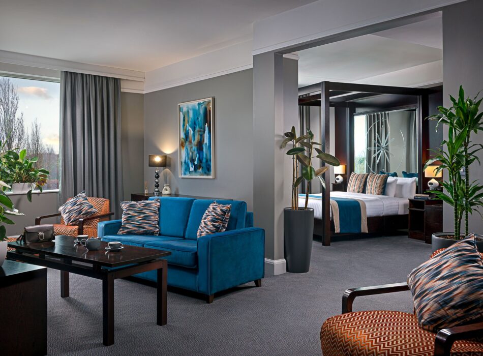 Spacious contemporary hotel suite with a separate bedroom and living area, featuring a blue sofa, patterned armchairs, modern artwork and indoor plants.