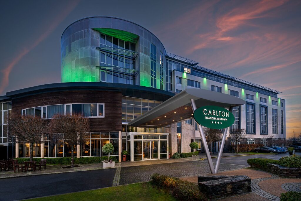 Exterior of the four-star Carlton Hotel Blanchardstown at dusk, with its modern entrance and curved façade illuminated by green lighting against a colourful evening sky.
