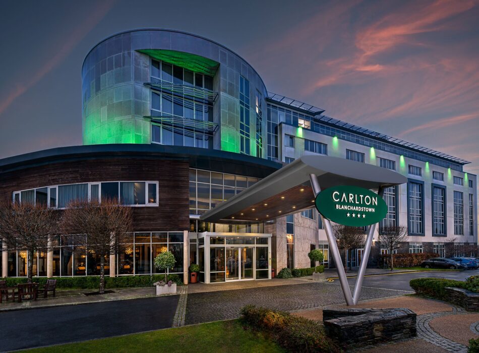 Exterior of the four-star Carlton Hotel Blanchardstown at dusk, with its modern entrance and curved façade illuminated by green lighting against a colourful evening sky.
