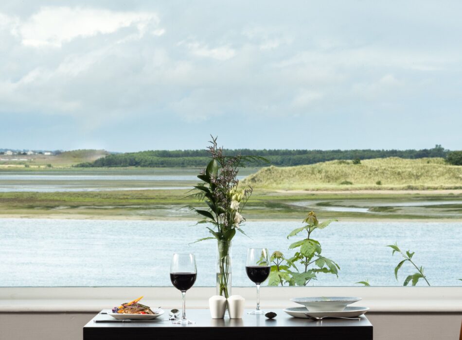 Dining table set beside a large panoramic window, overlooking calm coastal waters, sand dunes and the surrounding shoreline.