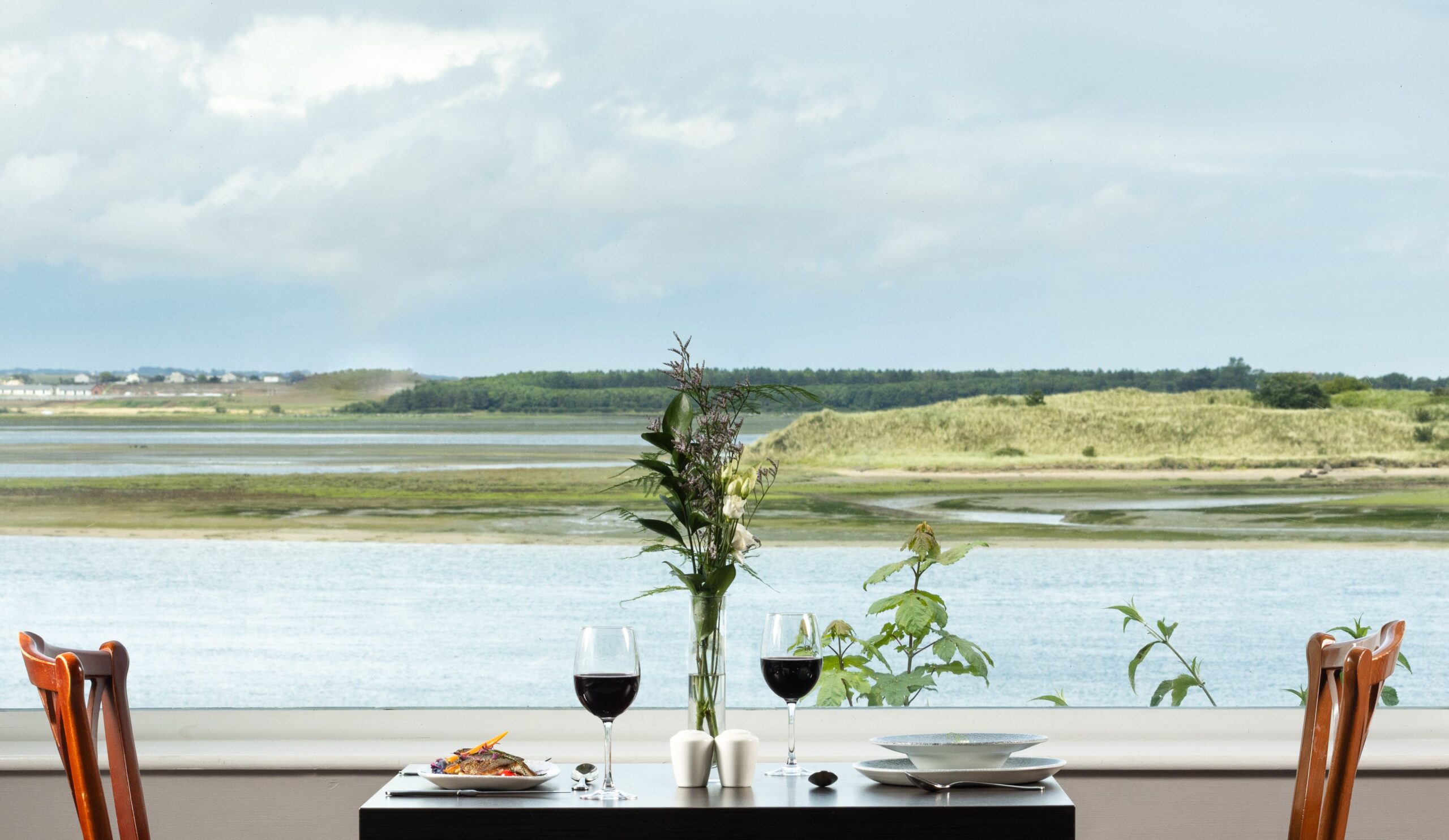 Dining table set beside a large panoramic window, overlooking calm coastal waters, sand dunes and the surrounding shoreline.