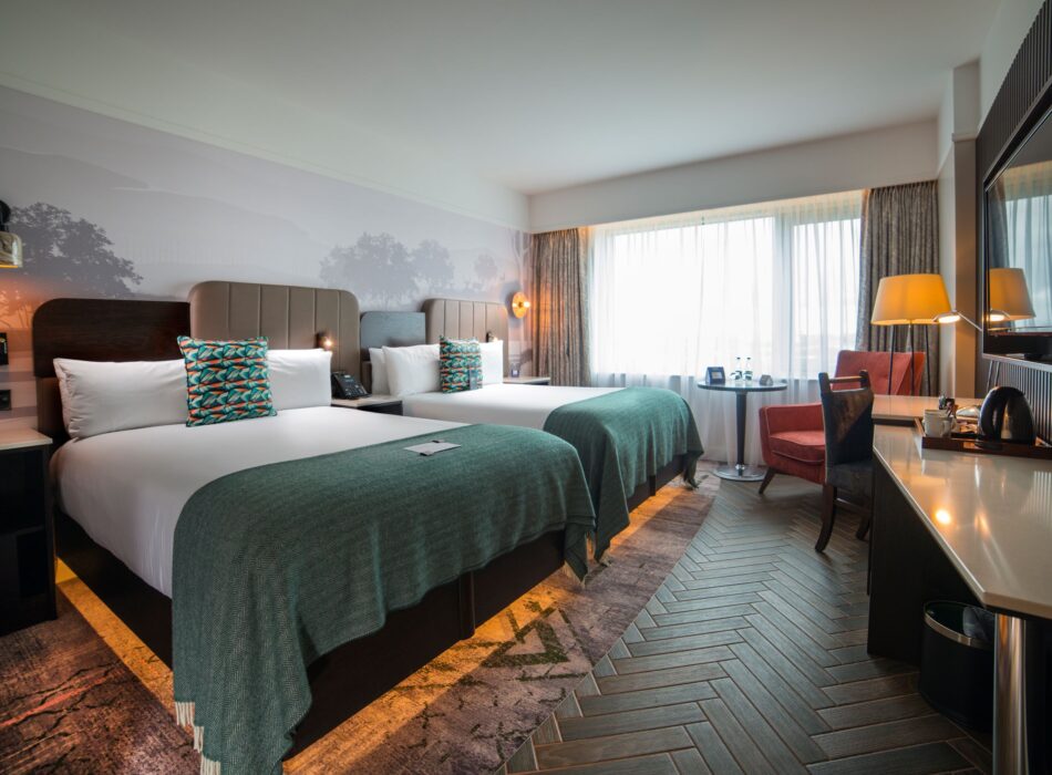 A spacious and stylishly designed hotel room containing two twin beds made up with crisp white linens, colorful patterned accent pillows, and dark teal knit throws. The main accent wall behind the beds is covered in a soft grey mural depicting a silhouette of trees. To the right, a long vanity console with a white countertop houses a television and a coffee station. The flooring shifts from a colorful patterned carpet under the beds to a dark grey herringbone tile pattern that leads toward a bright window framed by sheer curtains and an orange armchair.