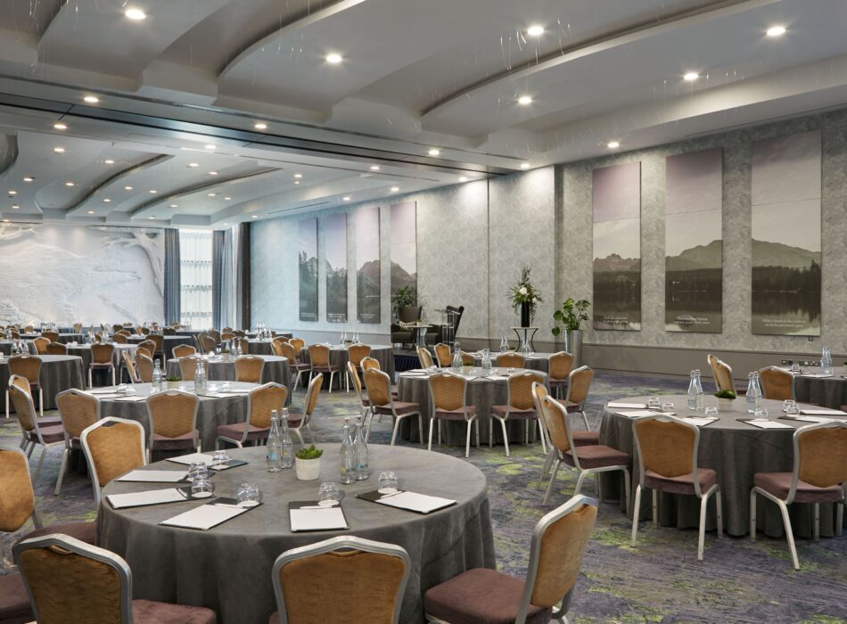 A sprawling, professionally styled ballroom configured for a large corporate function or meeting. Multiple round tables are covered with charcoal grey tablecloths and surrounded by brown fabric-backed dining chairs. Each table setting includes notebooks, pens, water bottles, and glasses. The expansive room features a striking modern ceiling with white, wave-like architectural panels and recessed spotlights. Long, vertical artwork panels depicting serene landscapes decorate the grey walls, and a large textured accent wall is visible at the far end of the room.