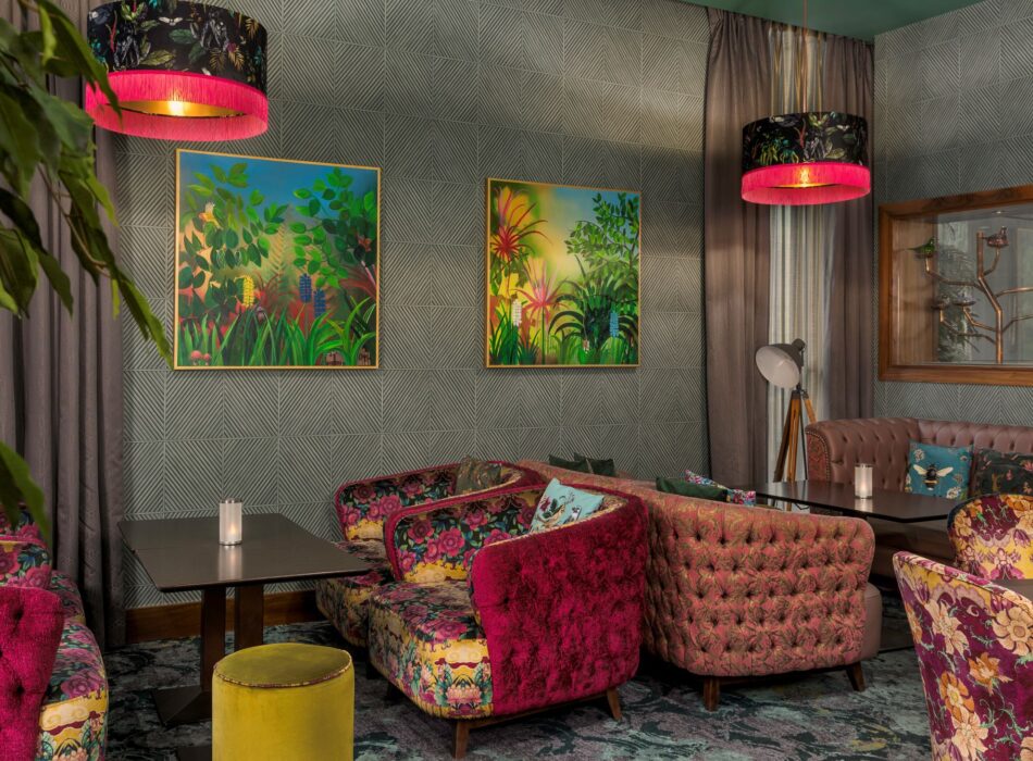A chic, maximalist seating area or lounge space. The walls are covered in a grey geometric-patterned wallpaper and adorned with two square paintings depicting dense, colorful tropical jungles. Tufted and smooth velvet armchairs are arranged around low black coffee tables, featuring various fabric designs including magenta pink floral print, dusty pink tufting, and purple floral upholstery. Two large drum pendant lights hang from above, showcasing a botanical print on the exterior and a bright, hot pink fringed trim along the bottom edge.