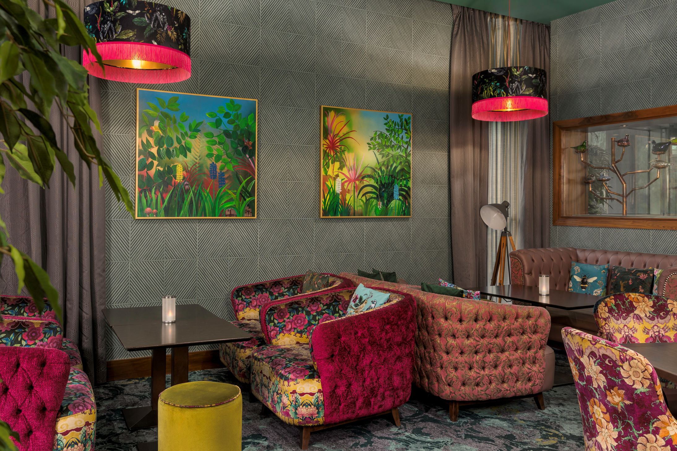 A chic, maximalist seating area or lounge space. The walls are covered in a grey geometric-patterned wallpaper and adorned with two square paintings depicting dense, colorful tropical jungles. Tufted and smooth velvet armchairs are arranged around low black coffee tables, featuring various fabric designs including magenta pink floral print, dusty pink tufting, and purple floral upholstery. Two large drum pendant lights hang from above, showcasing a botanical print on the exterior and a bright, hot pink fringed trim along the bottom edge.