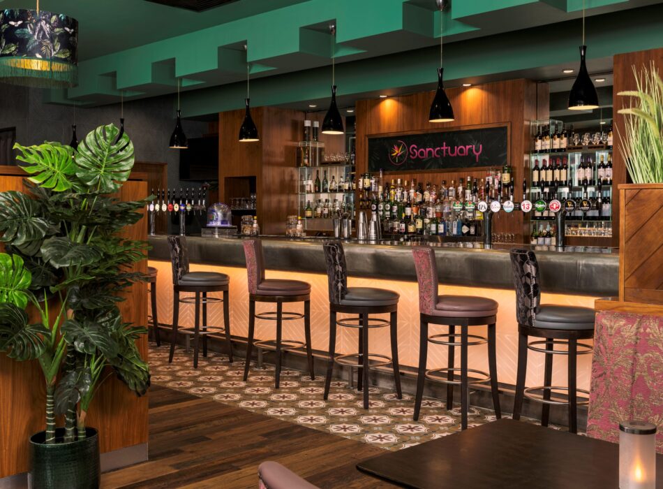 A chic hotel lounge bar with a polished counter lined with several high-backed, leather-upholstered barstools. The lower front of the bar glows with warm, amber geometric backlighting. Behind the counter, a wood-paneled wall displays a black sign reading "Sanctuary" in red script next to rows of liquor and spirit bottles. The room features a distinctive emerald green recessed ceiling with black pendant lights, a dark wood-plank floor with a patterned tile accent strip, and a large faux monstera plant in the left foreground.