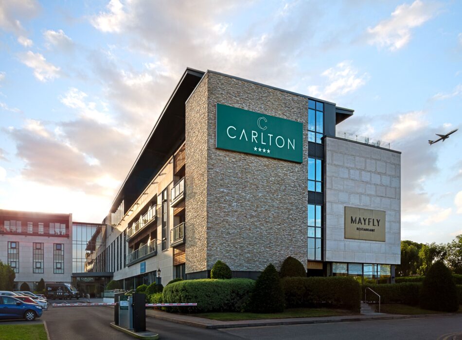 Exterior of the Carlton Hotel near Dublin Airport at sunset, showing the modern four-star hotel building and Mayfly Restaurant, with an aeroplane flying overhead.