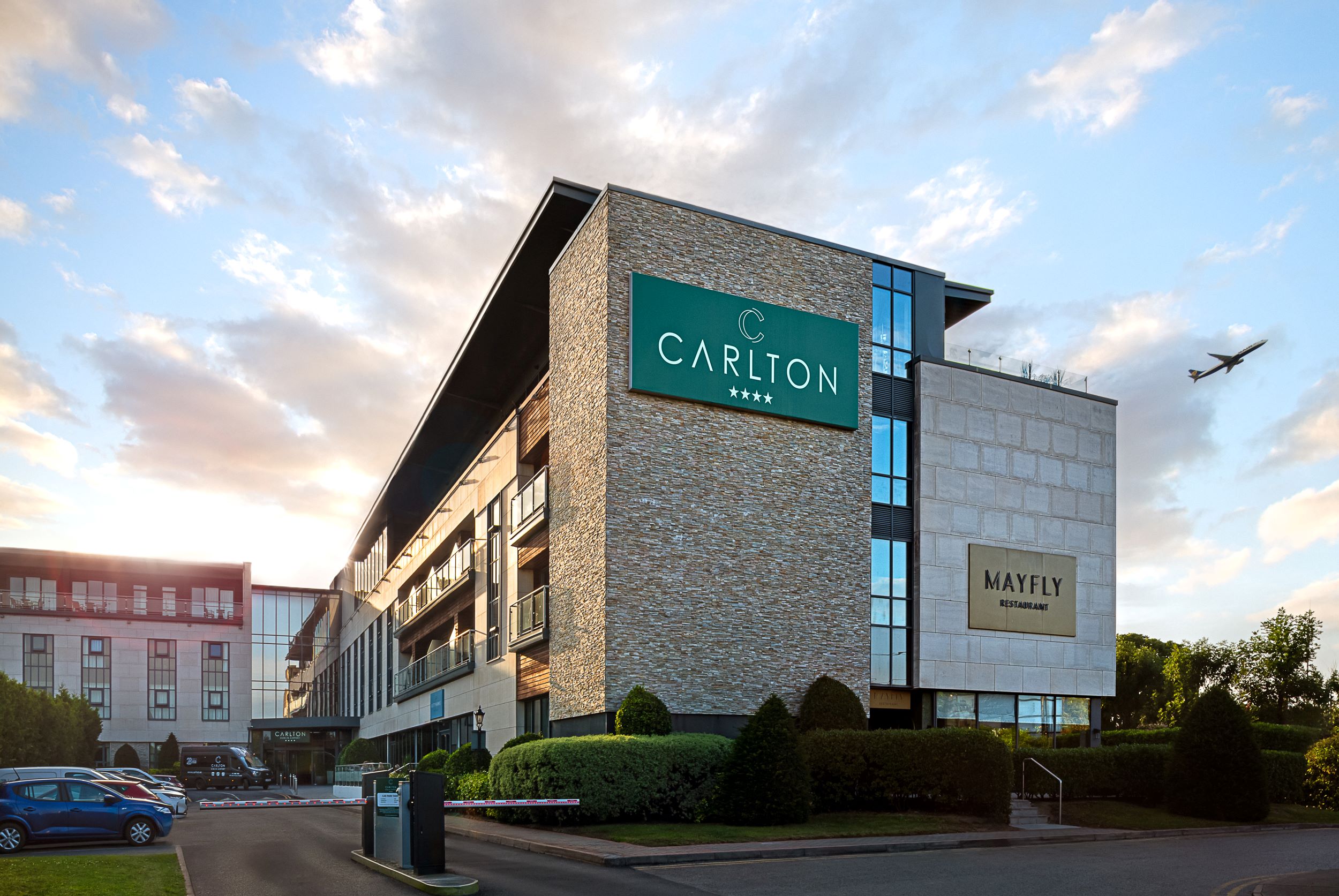 Exterior of the Carlton Hotel near Dublin Airport at sunset, showing the modern four-star hotel building and Mayfly Restaurant, with an aeroplane flying overhead.