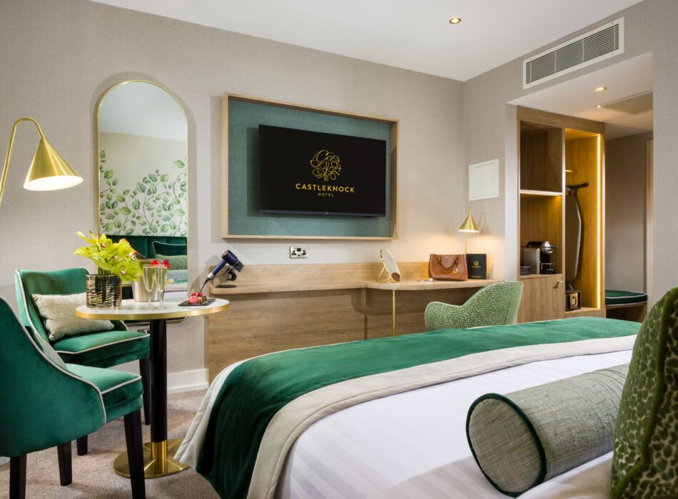 Interior view of a luxury hotel room at Castleknock Hotel. The foreground shows a bed with white linens and a green velvet throw. The room includes beige walls, light wood furniture, a mounted TV, two emerald green velvet chairs with a round table holding champagne, a tall arched mirror, and a built-in wardrobe with warm accent lighting.