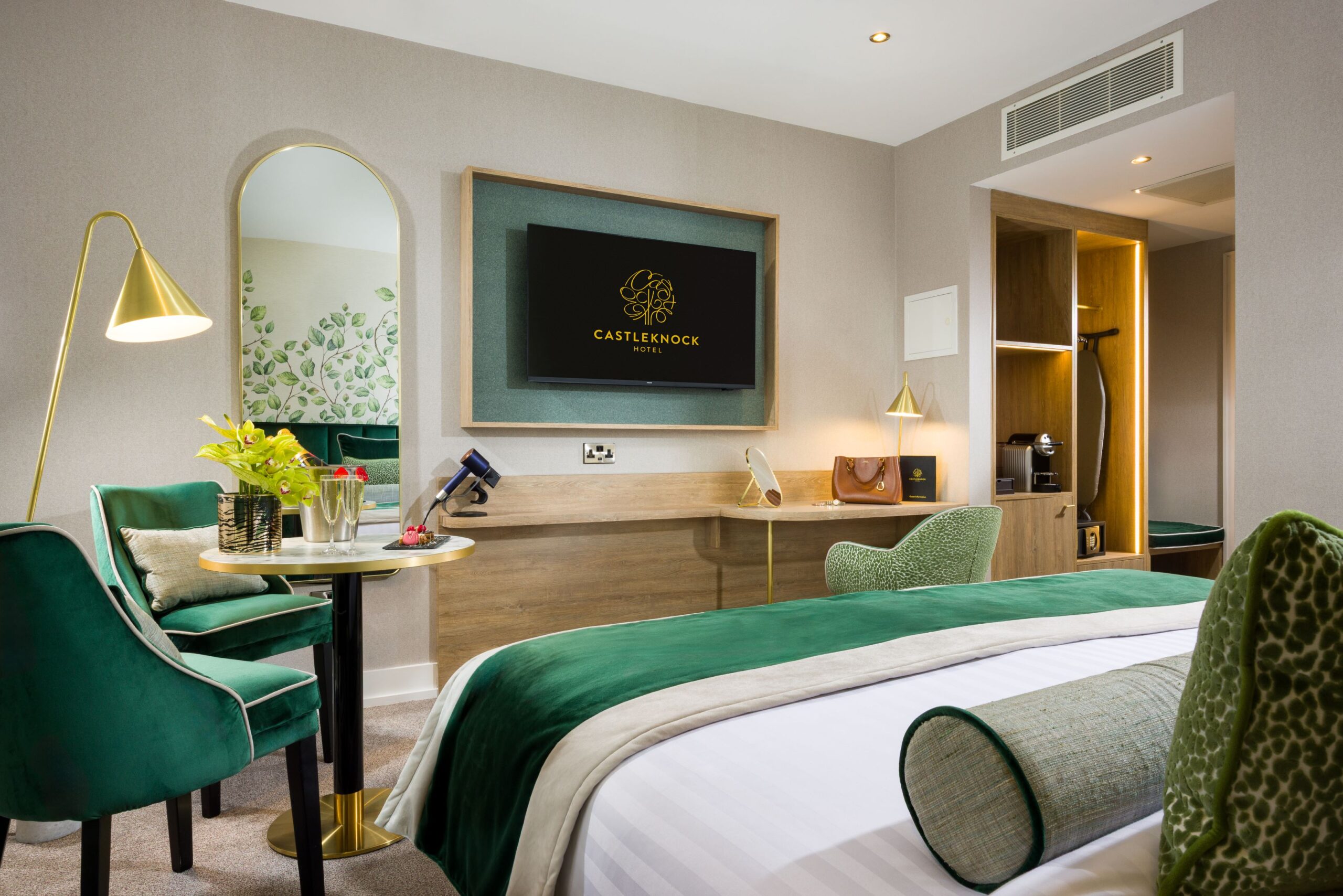 Interior view of a luxury hotel room at Castleknock Hotel. The foreground shows a bed with white linens and a green velvet throw. The room includes beige walls, light wood furniture, a mounted TV, two emerald green velvet chairs with a round table holding champagne, a tall arched mirror, and a built-in wardrobe with warm accent lighting.