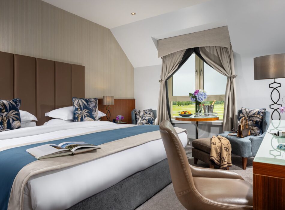 A bright, upscale hotel room featuring a large bed with a tall, brown tufted headboard, tropical-patterned pillows, and a teal blue bed runner. An open magazine rests on the bed. In the foreground, a brown leather office chair faces a wooden vanity desk. In the background, a small seating area with two patterned blue armchairs, a side table holding a wine bucket, and draped curtains frames a window overlooking a green golf course.