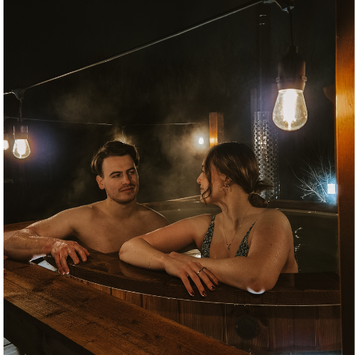 A couple relaxing together in an outdoor wooden hot tub at night, surrounded by steam and warm hanging lights at Hidden Acres.