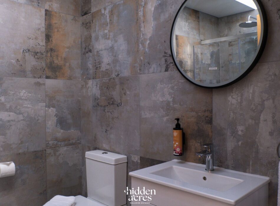 Modern bathroom at Hidden Acres featuring stone-effect tiled walls, a white sink and toilet, and a round mirror reflecting a walk-in shower.