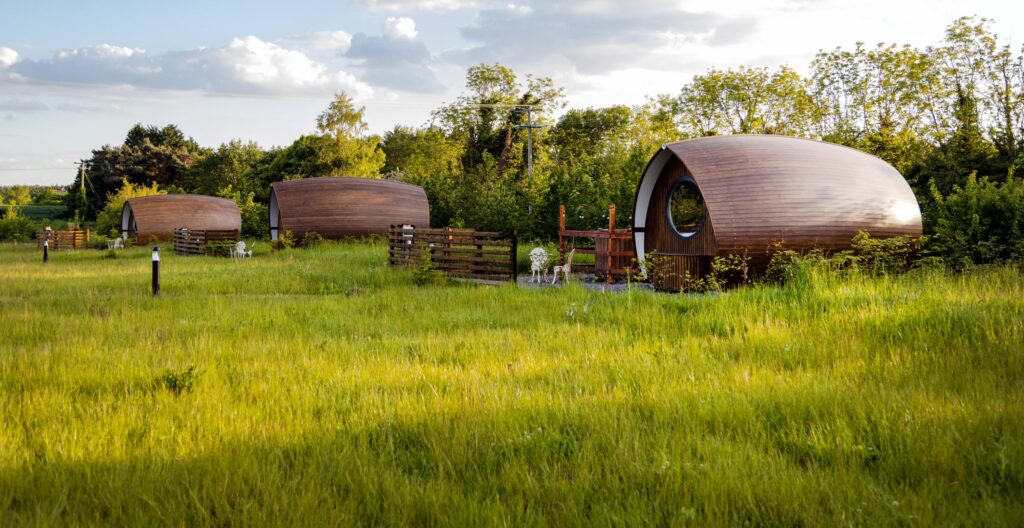 Three wooden glamping pods set among long grass and mature trees at Hidden Acres. Each pod has a small private outdoor seating area, creating a peaceful countryside accommodation setting surrounded by greenery.