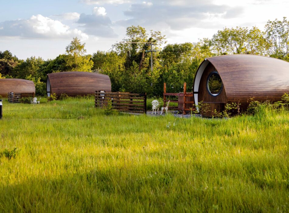 Three wooden glamping pods set among long grass and mature trees at Hidden Acres. Each pod has a small private outdoor seating area, creating a peaceful countryside accommodation setting surrounded by greenery.