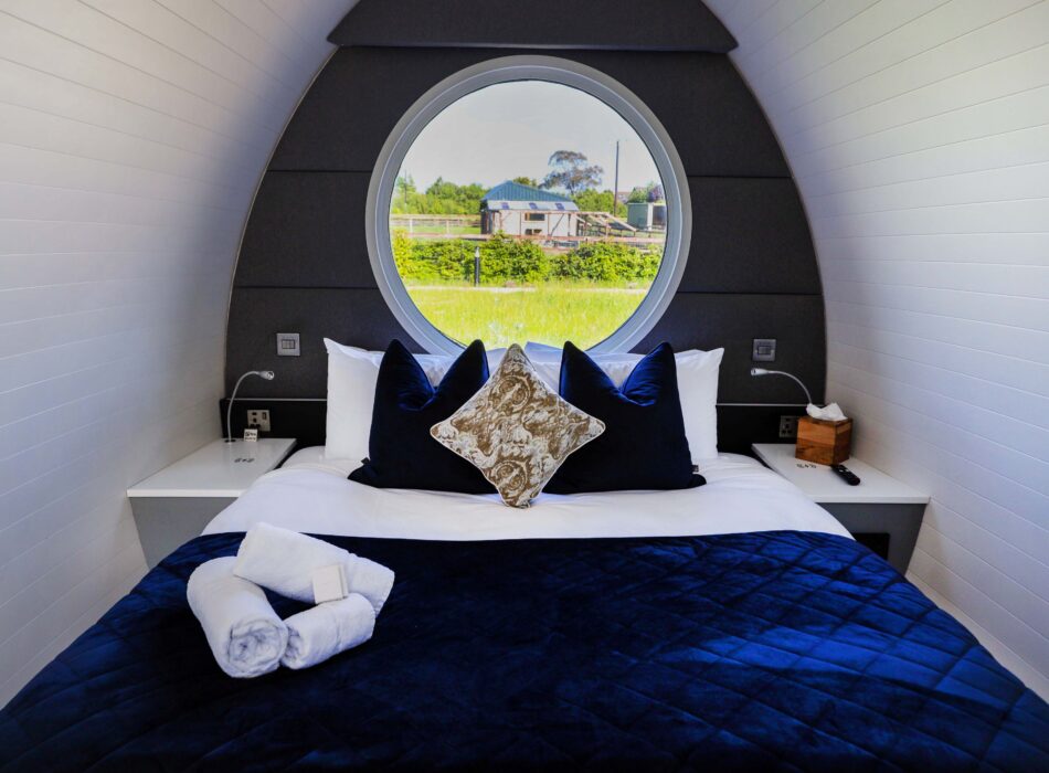 Stylish interior of a glamping pod at Hidden Acres in Malahide, featuring a neatly made double bed with deep blue bedding and decorative cushions. A large circular window above the bed frames a view of the surrounding green countryside.