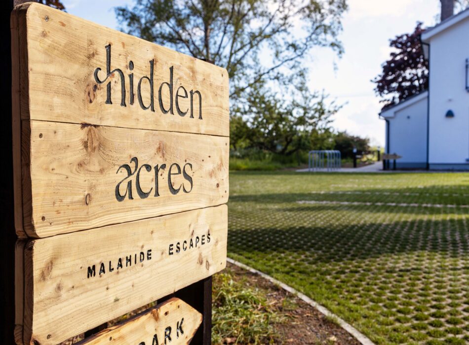 Close-up of a rustic wooden Hidden Acres sign at the entrance to the Malahide accommodation, with a modern white building, lawn and mature trees in the background.