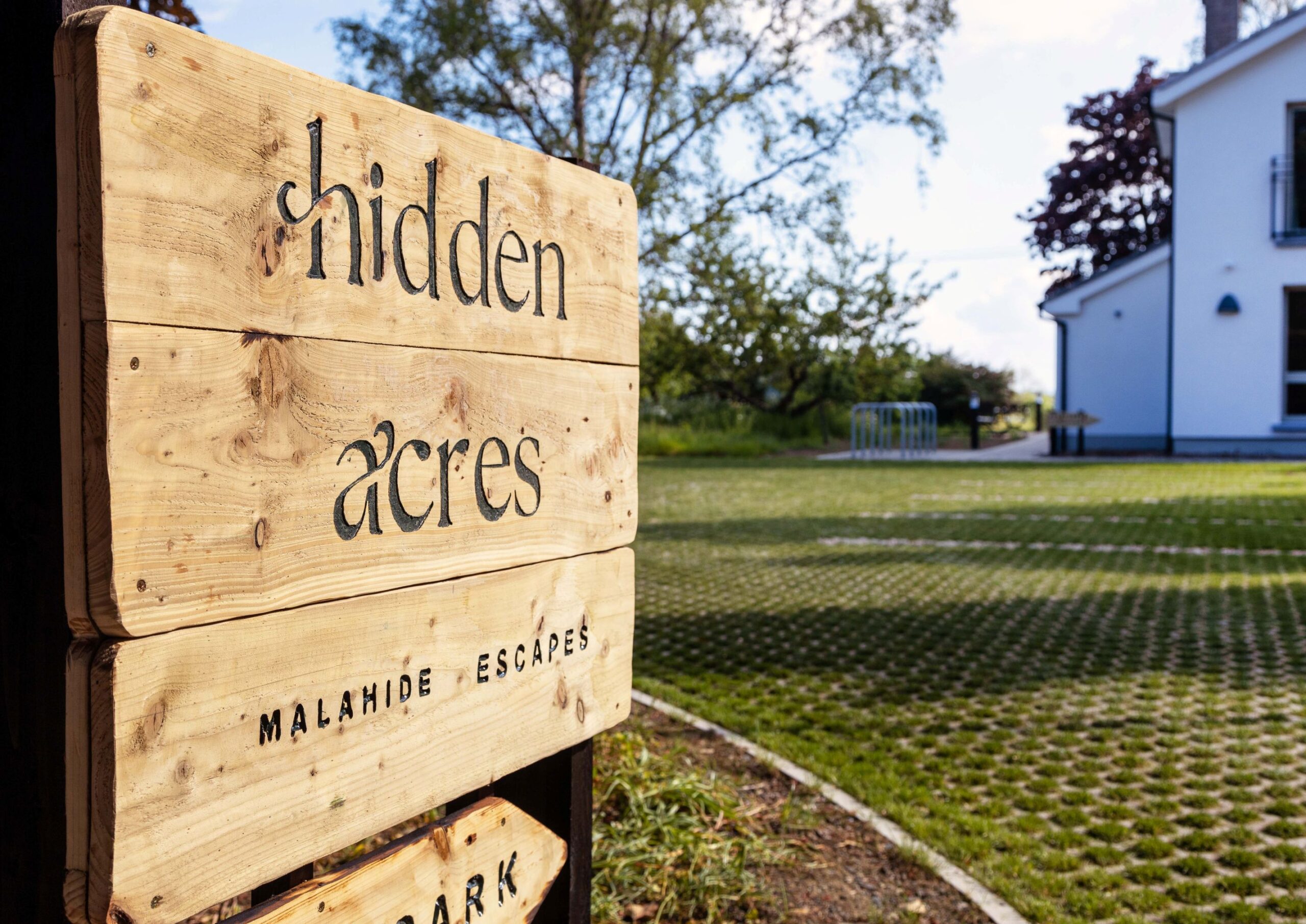 Close-up of a rustic wooden Hidden Acres sign at the entrance to the Malahide accommodation, with a modern white building, lawn and mature trees in the background.