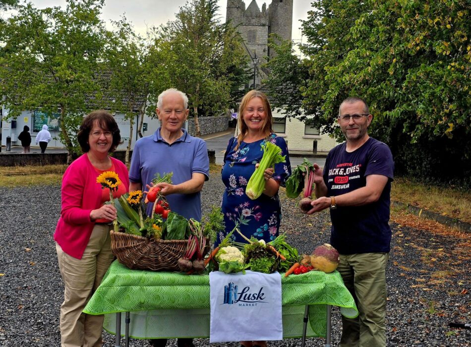 Four members of the Lusk community stand behind a colourful display of locally grown vegetables and flowers promoting Lusk Market, with the historic church tower visible in the background.