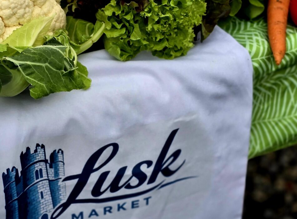 A close-up shot of local garden produce neatly arranged at an outdoor stall. A white fabric banner draping over the table prominently displays the "Lusk Market" logo in dark blue cursive script next to an illustration of a historic stone tower. Piled above it are various fresh vegetables, including a crisp head of white cauliflower with green leaves, a bunch of red-leaf and green curly lettuce, bright orange carrots, a bulb of fresh garlic, and green spring onions.