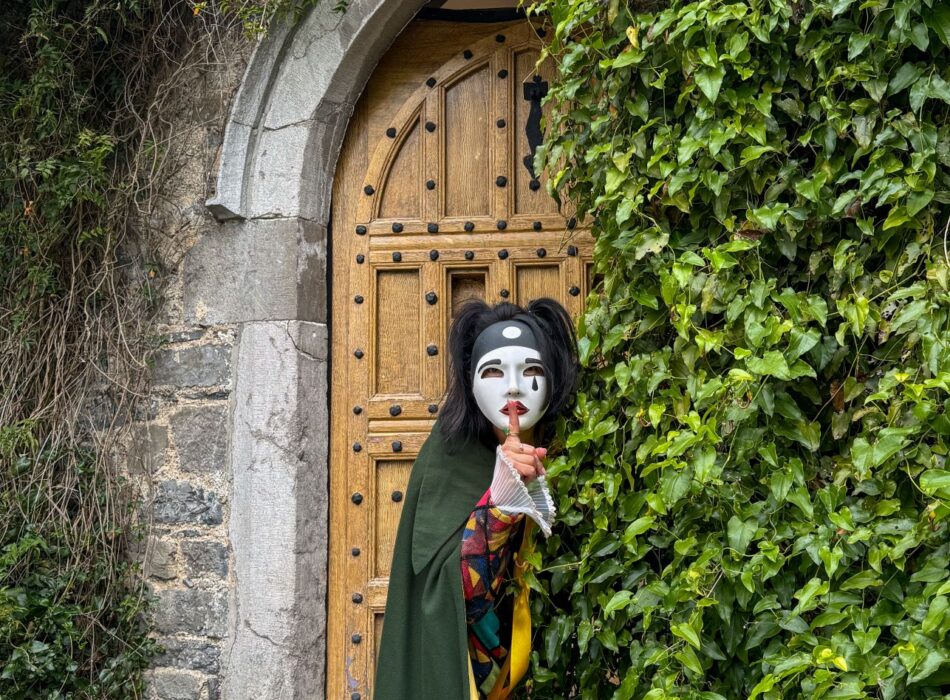 A person in a white mask, green cape, and colorful costume holds a finger to their lips while peeking out from ivy next to an arched wooden door.