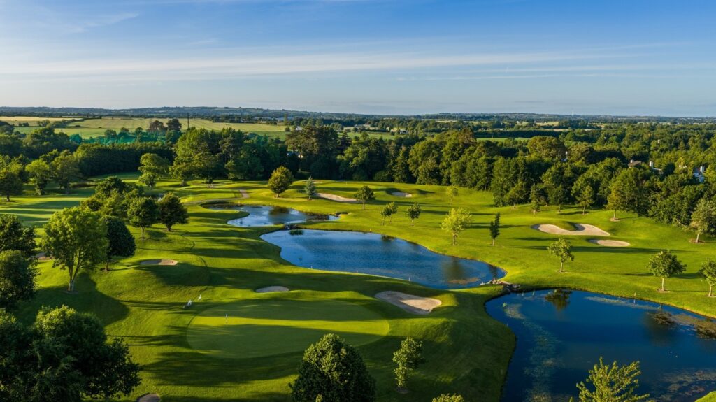 A high-angle aerial photograph capturing an expansive, rolling golf course illuminated by soft sunlight. The meticulously manicured green in the foreground is surrounded by rolling hills, strategic sand bunkers, and three interconnected blue lakes reflecting the sky. Scattered trees cast long afternoon shadows across the fairways, blending into a dense background of mature forests and distant, hazy green countryside beneath a light blue horizon. 