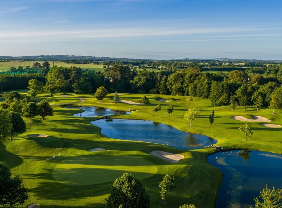 A high-angle aerial photograph capturing an expansive, rolling golf course illuminated by soft sunlight. The meticulously manicured green in the foreground is surrounded by rolling hills, strategic sand bunkers, and three interconnected blue lakes reflecting the sky. Scattered trees cast long afternoon shadows across the fairways, blending into a dense background of mature forests and distant, hazy green countryside beneath a light blue horizon.