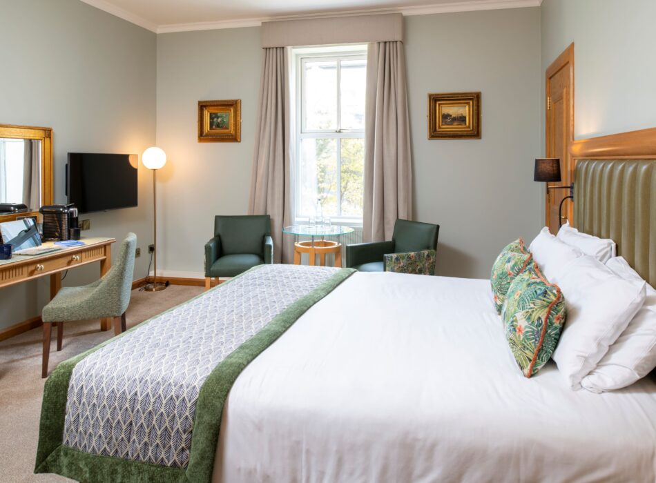 A bright hotel room with a large bed dressed in crisp white linens, botanical print accent pillows, and a textured green bed runner. The bed features a high, vertically paneled sage green leather headboard. To the left, a light wood vanity desk holds a laptop, a coffee machine, and a vanity mirror beneath a wall-mounted flat-screen TV. In the background, two dark green leather armchairs flank a small glass table in front of a window dressed with heavy beige curtains.