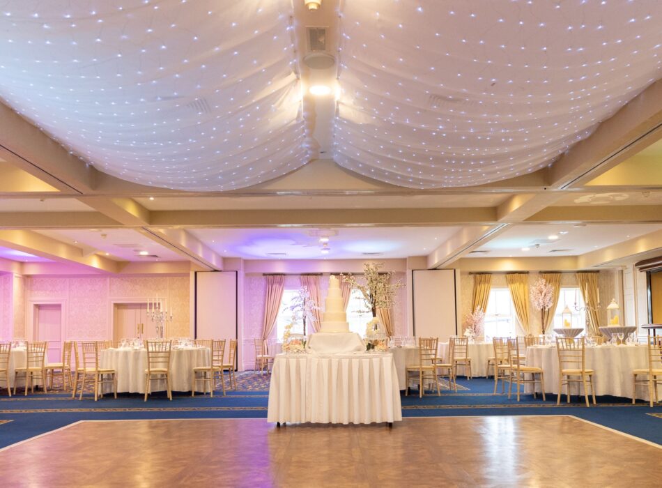 A wide interior shot of a spacious banquet hall decorated for a wedding. In the foreground, a glossy wooden dance floor is flanked by a blue patterned carpet. A central display table holds a tall, white multi-tiered wedding cake flanked by decorative white blossom trees. Round tables dressed in white tablecloths and paired with gold Chiavari chairs fill the room. The ceiling is elegantly draped with white fabric billows embedded with twinkling white fairy lights, while ambient purple lighting glows along the side walls.
