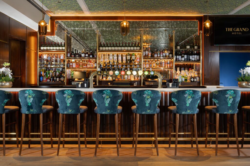 > Stylish hotel bar with teal upholstered stools, warm lighting and an extensive display of wines, spirits and draught beers behind the counter.
